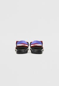 Sandales de sport avec une semelle noire, des sangles violettes, rouges et blanches, avec un design plat et des fermetures réglables pour un ajustement sécurisé.