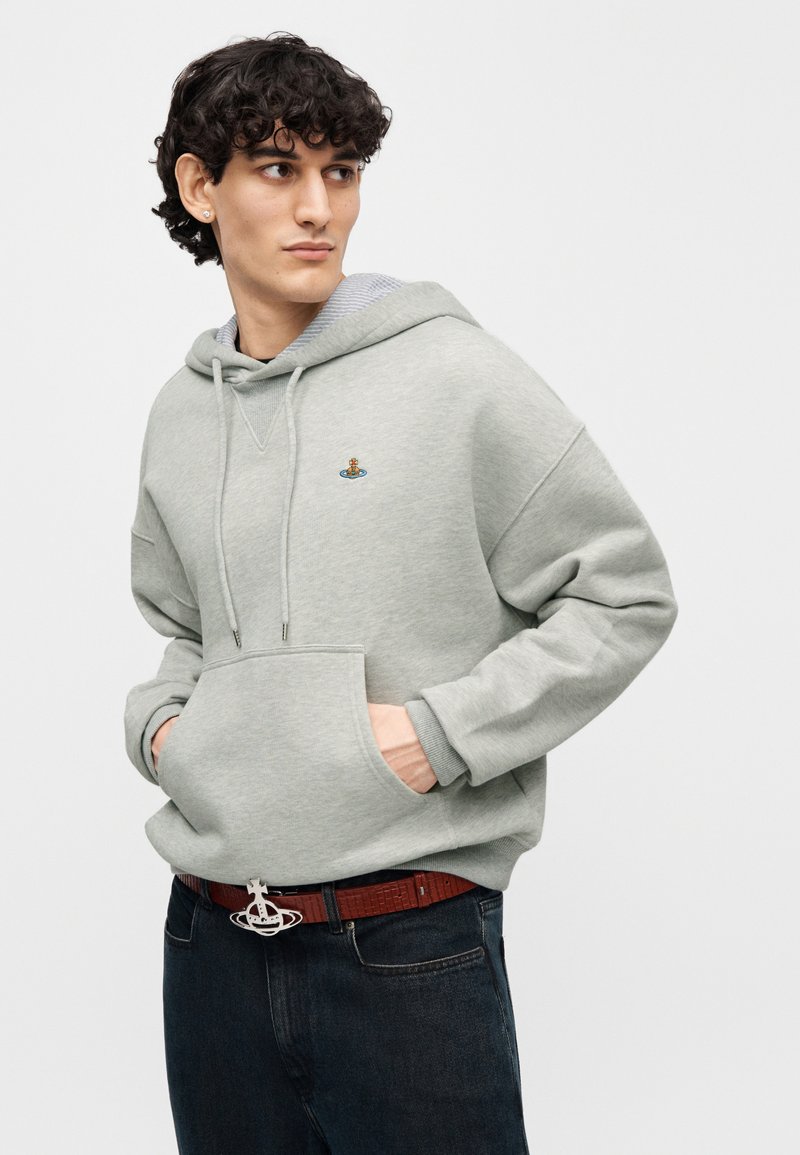 Sweat à capuche en coton gris clair avec poche frontale, capuche à cordon et motif brodé, associé à un jean foncé et une ceinture marron.