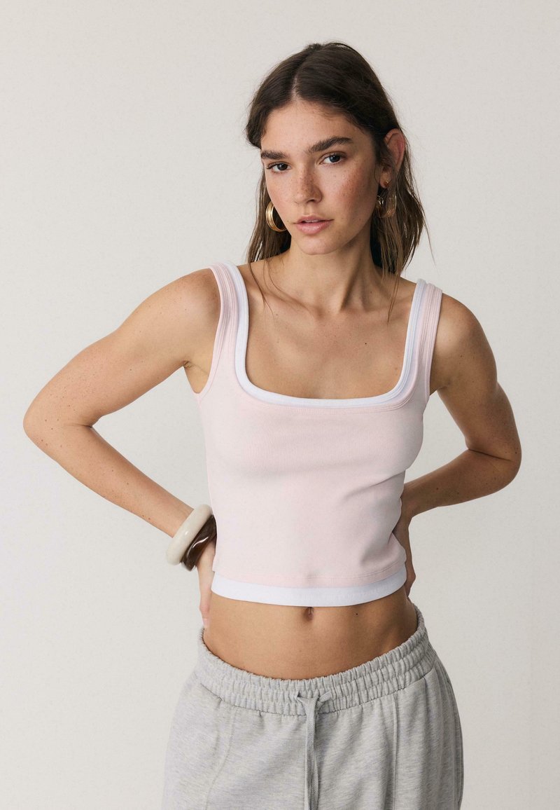 Jeune femme aux longs cheveux bruns portant un haut court sans manches rose pâle, un pantalon de survêtement gris, des boucles d'oreilles créoles et des bracelets, posant les mains sur les hanches.