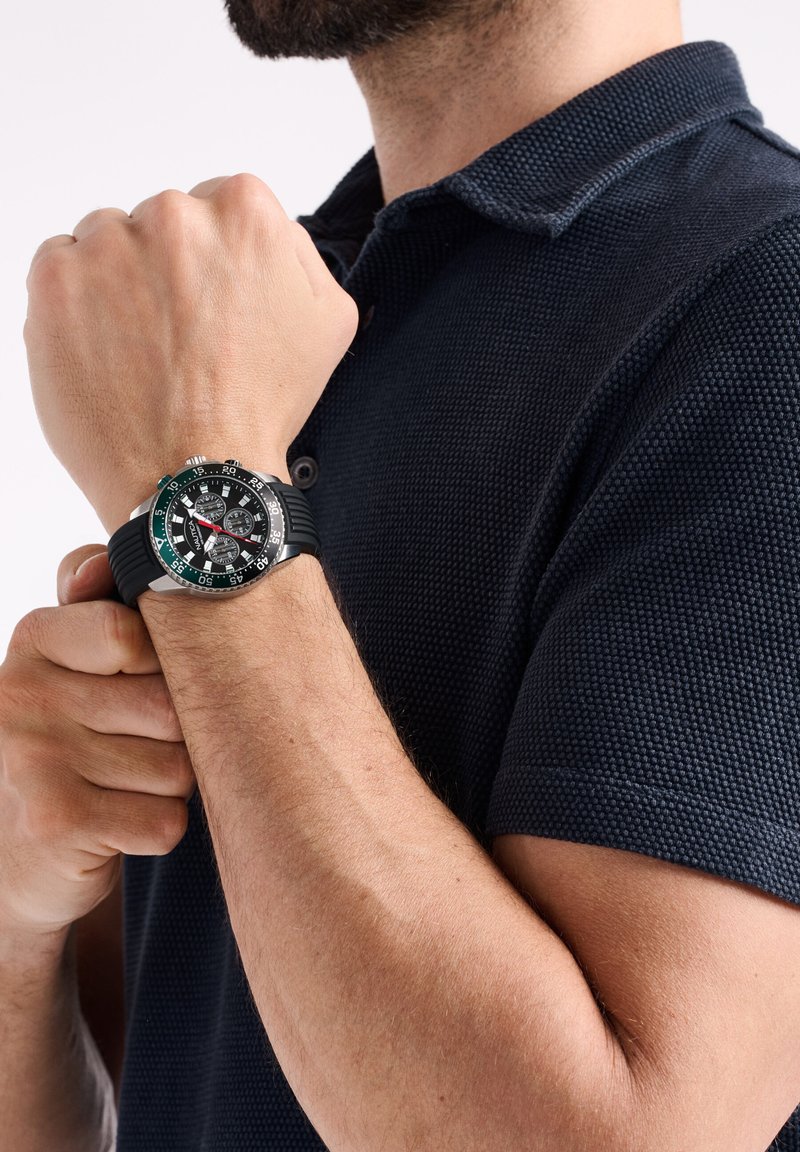 Melna gumijas josta pulkstenis ar apaļu sudraba korpusu, melnu ciparnīcu, baltām atzīmēm un zaļiem akcentiem. Valkāts uz plaukstas, kur redzama āda.