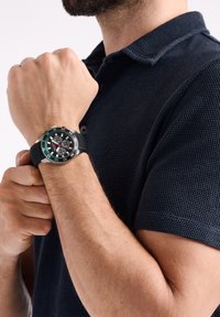 Uomo che indossa una polo di tessuto blu scuro mentre regola un orologio cronografo nero con lunetta argentata al polso sinistro.