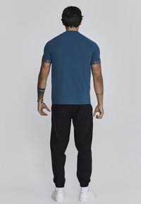 Camisa azul de manga corta con mangas raglán, que presenta acentos de logo en los puños; combinada con pantalones negros elásticos con puños y zapatillas blancas.