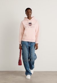 Roze hoodie met logo, grote voorkeurszak, gecombineerd met losse blauwe jeans en witte sneakers. Model houdt een kleine rode handtas vast.