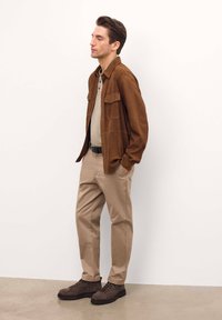 Jachetă din suede maro peste un polo bej, pantaloni bej și cizme din piele maro închis cu șireturi. Textură netedă, cu o croială lejeră și două buzunare la piept.