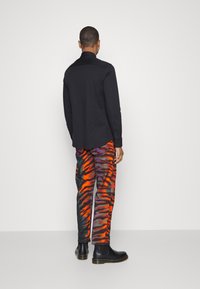 Maglietta nera a maniche lunghe abbinata a pantaloni dai motivi astratti colorati in arancione, viola e nero. L'individuo indossa stivaletti neri alla caviglia.