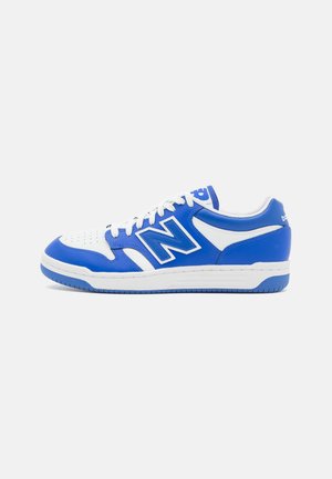 Baskets basses de sport en bleu et blanc avec des lacets blancs et un grand logo bleu "N" sur le côté, sur un fond clair uni.