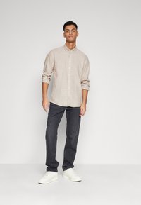 Camicia button-up beige chiaro con maniche arrotolate, abbinata a jeans grigi scuri e sneakers bianche, in piedi contro uno sfondo semplice.