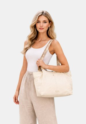 Jonge vrouw met golvend blond haar die een wit tanktop en beige broek draagt, met een grote crèmekleurige tas over haar schouder.