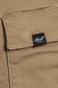 Beige Baumwollstoff mit einer Taschenklappe; sichtbar genäht. Schwarzes Etikett mit dem "Reell"-Logo, auf die Tasche genäht.