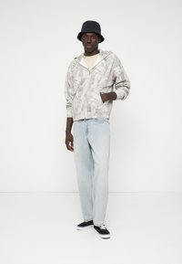 Grijze zip-up hoodie met een bladerenpatroon, gecombineerd met lichtblauwe jeans. Een zwarte bucket hat en zwarte sneakers maken de outfit compleet.