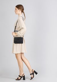 Černá krosbody taška s kruhovým logem, plochý design a nastavitelný popruh, v kombinaci s béžovými šaty a černými sandály na blokovém podpatku.