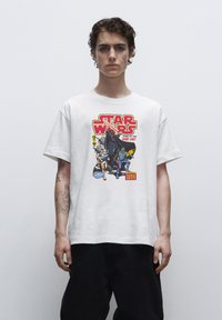 Bílý bavlněný tričko s barevným grafickým potiskem "Star Wars", který zobrazuje postavy a text. Krátké rukávy, volný střih, hladká textura.