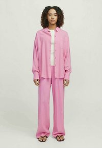 Roza prevelika srajca z gumbi in dolgimi rokavi, v kombinaciji z ujemajočimi se širokimi hlačami. Gladek material; manekenka stoji v natikačih.