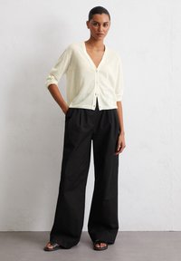 Helle gelbe Strickjacke mit V-Ausschnitt, Knopfriegel und langen Ärmeln, kombiniert mit weiten schwarzen Hosen und flachen Sandalen.