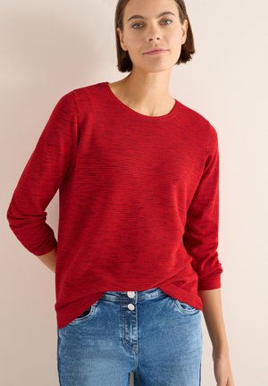 Rotes Langarmoberteil mit strukturiertem Streifenmuster, runder Ausschnitt und lässiger Passform, kombiniert mit blauen Denim-Jeans mit Knopfdetails.