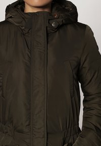 Doudoune vert olive avec capuche, dotée de boutons-pression, de poches latérales et d'une taille froncée pour une silhouette ajustée. Texture lisse et brillante.