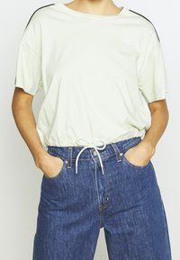 Camiseta de manga corta verde claro con cuello redondo, cordón elástico en el dobladillo y detalles negros en las mangas; combinada con unos vaqueros azules.