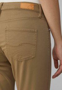 Khaki spodnie o gładkiej fakturze, z tylną kieszenią z zaokrąglonym przeszyciem i brązową skórzaną naszywką na pasku.
