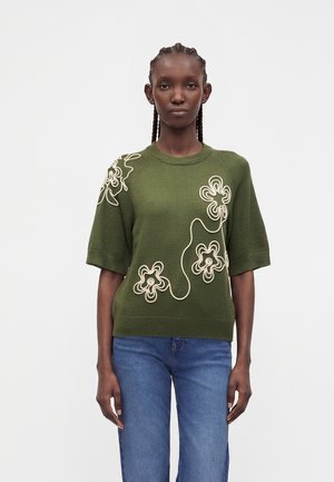 Femme aux cheveux tressés portant un pull manches courtes vert olive avec broderies florales blanches et un jean bleu, debout devant un fond uni.