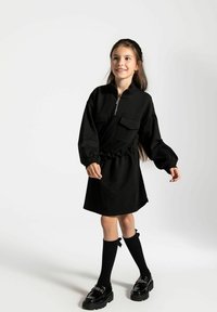 Czarna sukienka z kieszeniami, z pół-zamkiem przy dekolcie, elastycznym pasem, długimi rękawami, zestawiona z podkolanówkami i czarnymi butami. Gładka tekstura materiału.