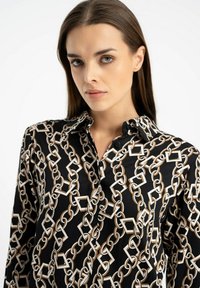 Czarna koszula z wzorem łańcucha w białym i beżowym kolorze, z kołnierzykiem, zapięciem na guziki oraz długimi rękawami. Gładka faktura materiału.