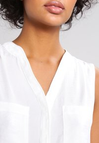 Gros plan sur le cou et les lèvres d'une personne portant un débardeur blanc à col en V avec des poches sur la poitrine.