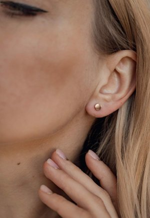 Goldfarbene Steckerohrringe mit einer glatten, runden Oberfläche, die am Ohrläppchen befestigt sind. Nahaufnahme eines Ohrs und einer Hand, die den Hals berührt.