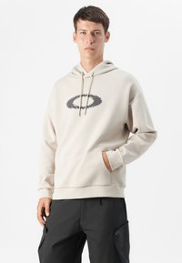Ung mand iført en beige hættetrøje med et logo, stående med den ene hånd i lommen, mod en ensfarvet hvid baggrund.