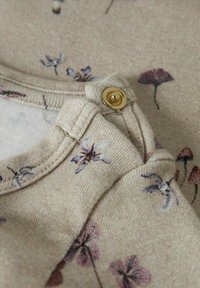 Primo piano di un tessuto beige con delicato motivo floreale viola e un bottone a pressione in ottone sul bordo piegato del capo.