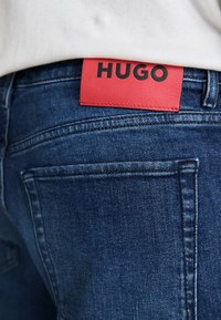 Dżinsy jeansowe w ciemnoniebieskim odcieniu z widoczną czerwoną skórzaną łatką z napisem „HUGO” na plecach. Posiadają standardowe kieszenie i detale szwów.