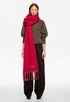 Kobieta nosząca duży, jaskraworóżowy szal z frędzlami, nałożony na brązowy, puszysty kardigan i luźne ciemne spodnie, stojąca na tle białego tła.