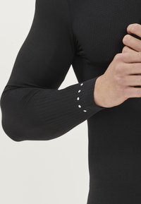 Svart långärmad skjorta med texturerad kropp och ribbad handled. Har fem små vita prickar längs manschetten. Materialet verkar töjbart och åtsittande.