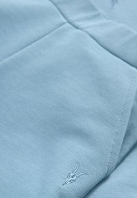 Ljusblå bomullsplagg med mjuk textur, prydd med en vit broderad antiloplogotyp. Sömmarna är snygga och tydligt definierade.