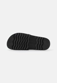 Semelle de chaussure noire avec un motif de bande de roulement ondulé, affichée à plat sur un fond gris clair.