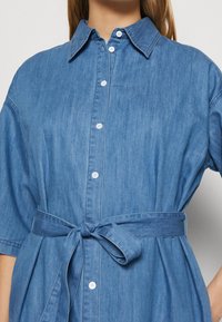 Jasnoniebieska sukienka koszulowa z denimu, z krótkimi rękawami, zapięciem na guziki i przewiązaną w talii szarfą. Gładka faktura, casualowy design i kołnierz.
