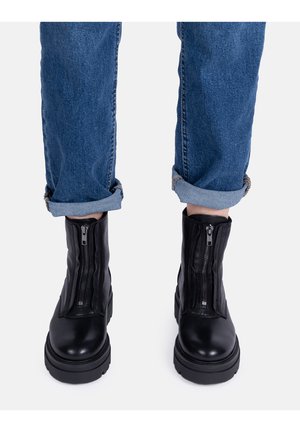 Eine Person trägt blaue, hochgekrempelte Jeans und schwarze Leder-Ankle-Boots mit Vorderreißverschluss und steht vor einem weißen Hintergrund.