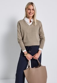 Beige V-neck sweater with ribbed cuffs, paired with navy pants. Holding a large taupe tote bag, featuring minimal hardware and smooth texture.