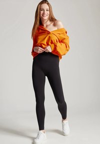 Pomarańczowy sweatshirt bez rękawów w zestawieniu z czarnymi legginsami; modelka ma na sobie białe trampki; materiał wydaje się miękki i elastyczny.