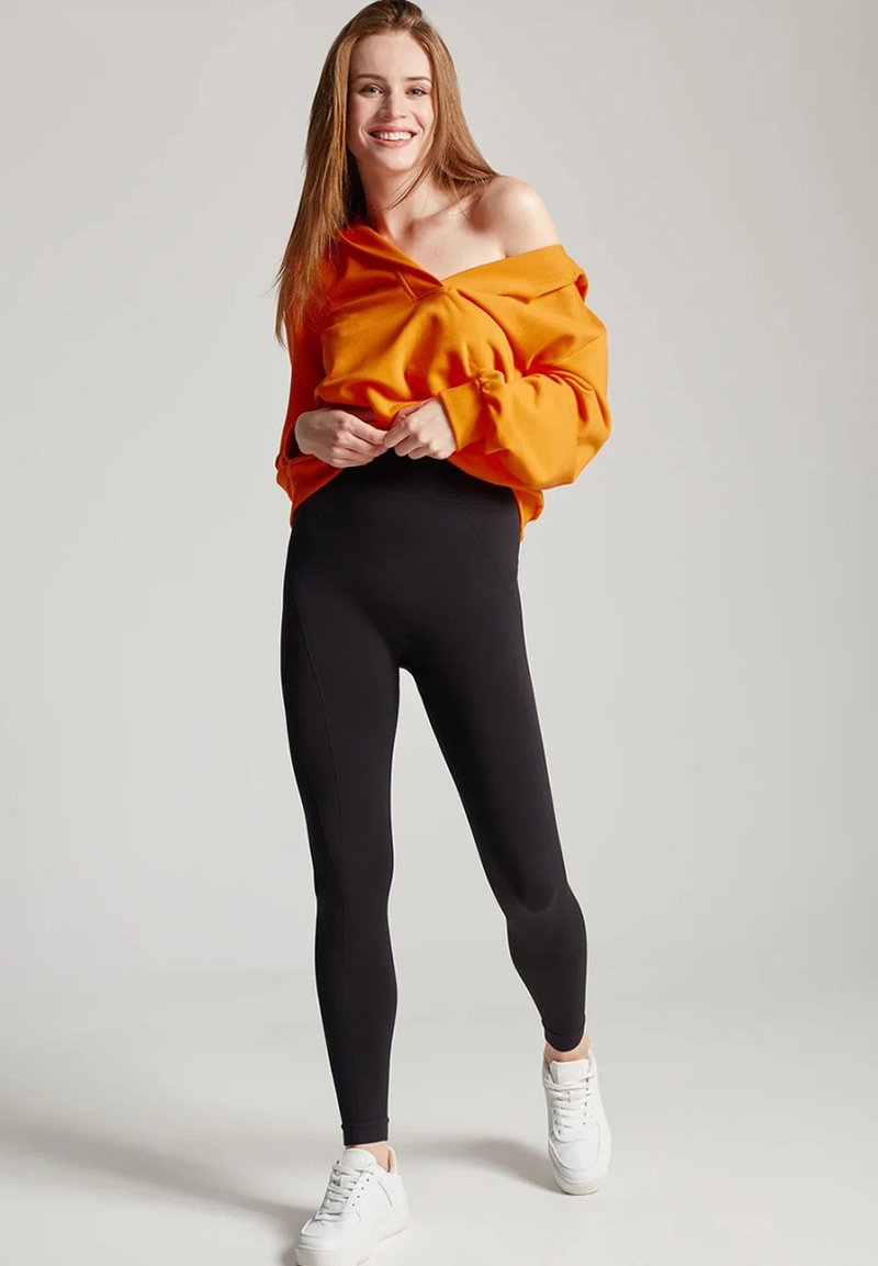 Pomarańczowy sweatshirt bez rękawów w zestawieniu z czarnymi legginsami; modelka ma na sobie białe trampki; materiał wydaje się miękki i elastyczny.