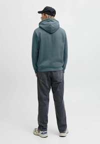 Kapuzensweatshirt in Türkis mit weichem Material, kombiniert mit dunkelgrauen Jeans. Das Outfit wird ergänzt durch sportliche Schuhe und eine schwarze Kappe.