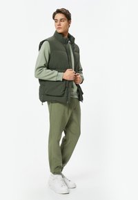 Chaleco acolchado en verde oscuro, combinado con una sudadera verde a juego y joggers. Zapatillas blancas completan el atuendo. Textura de tela suave.