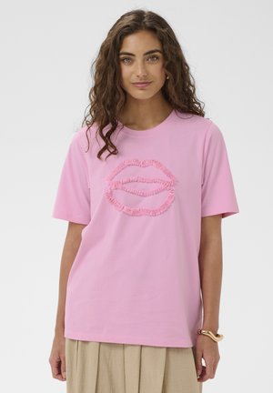 Göndör barna hajat viselő nő rózsaszín pólóban, amelyen egy textúrált ajak motívum látható, bézs szoknyában, egy egyszerű világos háttér előtt áll.