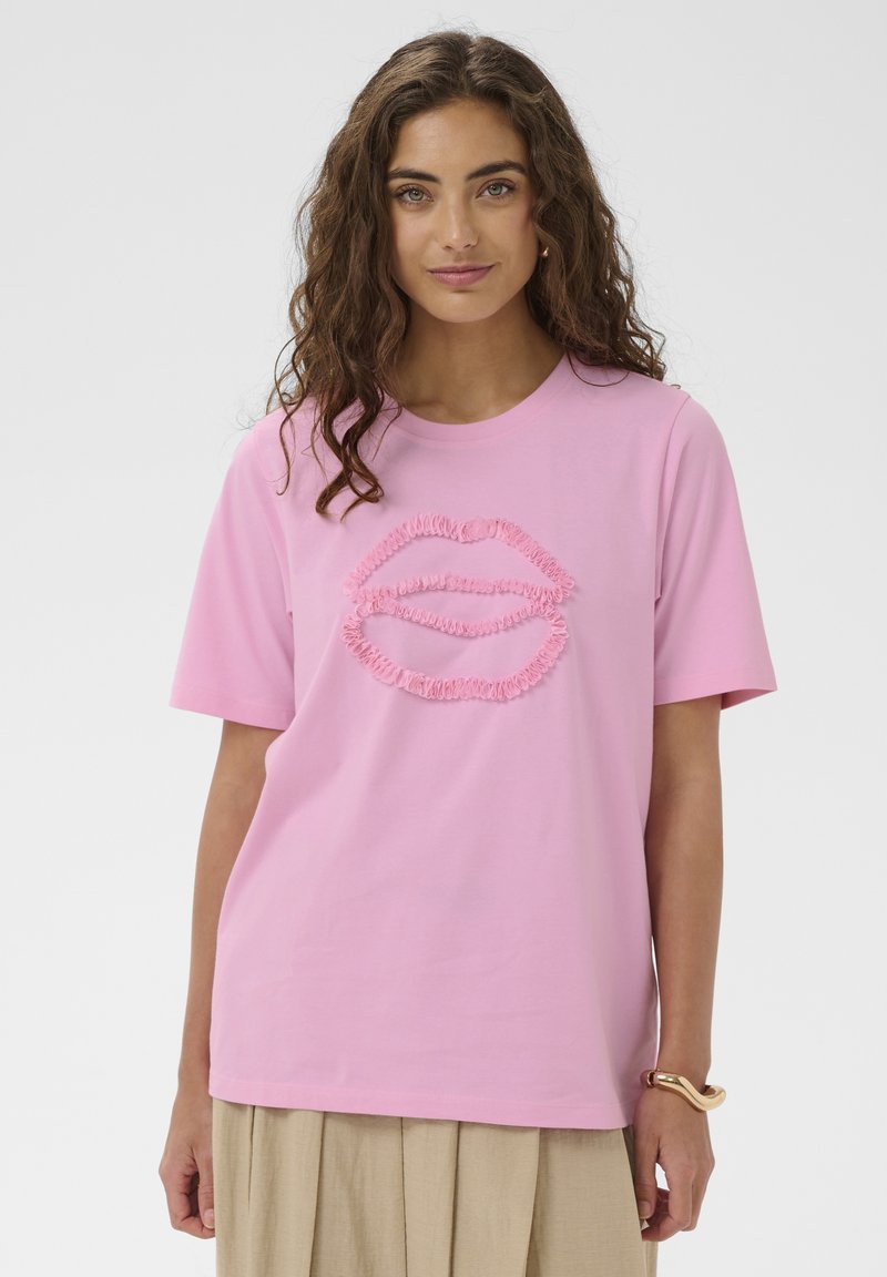 Donna con capelli ricci castani che indossa una maglietta rosa con un design labbra in rilievo e una gonna beige, in piedi davanti a uno sfondo chiaro e semplice.