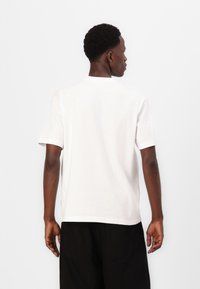 Weißes Baumwoll-T-Shirt mit kurzen Ärmeln und rundem Halsausschnitt, von hinten präsentiert, kombiniert mit schwarzen Hosen. Einfaches Design, ohne Muster.