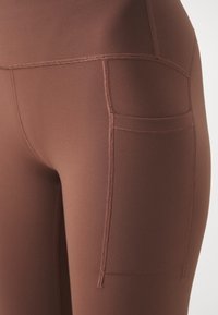 Bruna leggings med en slät textur, med en sidficka och kontrasterande sömmar för extra detalj. Tätsittande design.