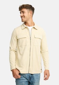 Ung mand med kort, krøllet hår, der smiler, iført en beige lynlås cordjakke over en hvid skjorte og blå jeans, stående op ad en hvid baggrund.