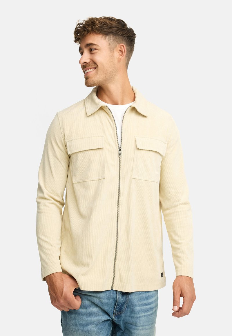Ung mand med kort, krøllet hår, der smiler, iført en beige lynlås cordjakke over en hvid skjorte og blå jeans, stående op ad en hvid baggrund.
