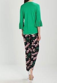 Haut en maille vert avec manches trois-quarts plissées, associé à un pantalon noir à motif floral avec des fleurs roses. Le mannequin porte des talons argentés.