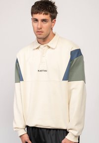 Jersey crema con cuello estructurado, con detalles en verde y azul en las mangas, y logotipo "KAOTIKO" en la zona del pecho. Textura suave.