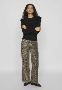 Melnas garas piedurknes blūze ar frillām, apvienota ar leopardu raksta platām biksēm un melniem dzīvokļiem. Neitrāls fons.